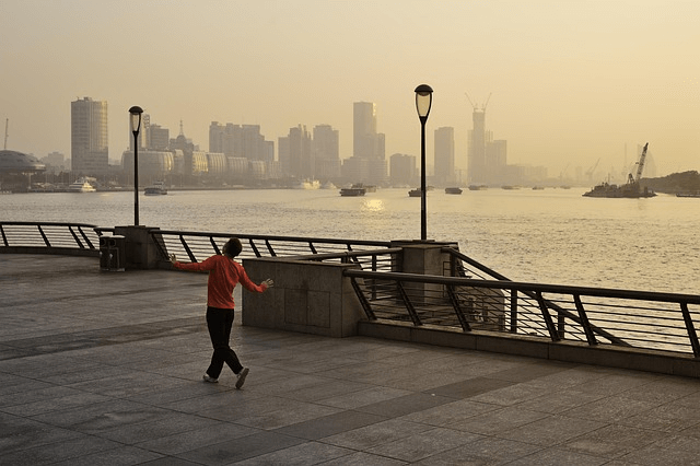 man in red hoodie is walking funnily on a promenade next to a river on hazy day with city skyline in the background