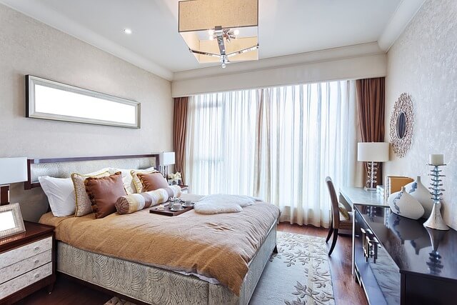 picture of beautiful hotel room in warm colors of light beige and shades of brown colors, kingsize bed on left with pillows of different shapes and colors and beige throw, on right chest of drawers and desk with a chair, lamps and with round mirror