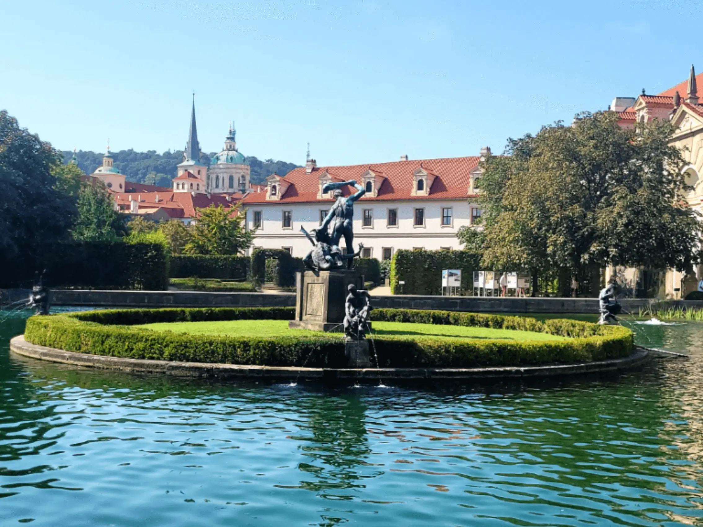 Waldstein Garden in Prague showing turquoise lake and green island with a statue in the middle with background of historic buildings